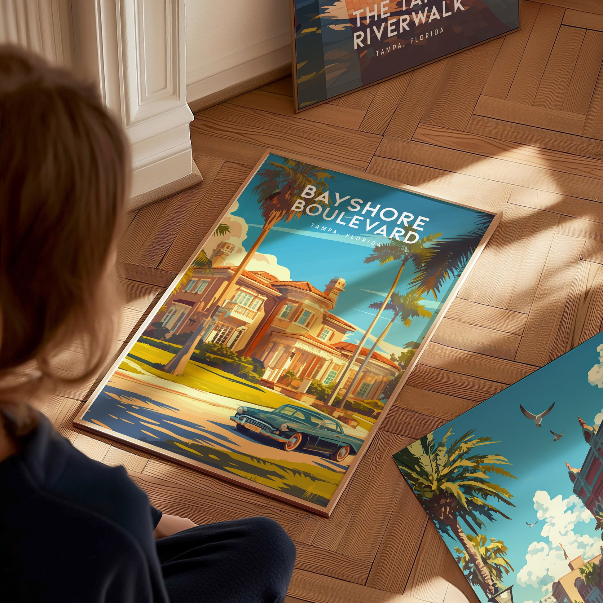 A person sits on a wooden floor looking at a colorful poster of Bayshore Boulevard, featuring a classic car and palm trees. Sunlight casts shadows across the scene.