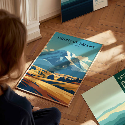 A person sits on the wooden floor, looking at a Mount St. Helens poster depicting a stylized landscape with a mountain, clouds, and fields. Other posters are partially visible nearby.
