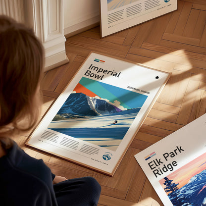 A person sits on a wooden floor, looking at a colorful travel print featuring a stylized mountain landscape with a skier in the foreground. The print is part of a collection showcasing various destinations, with text labels visible.