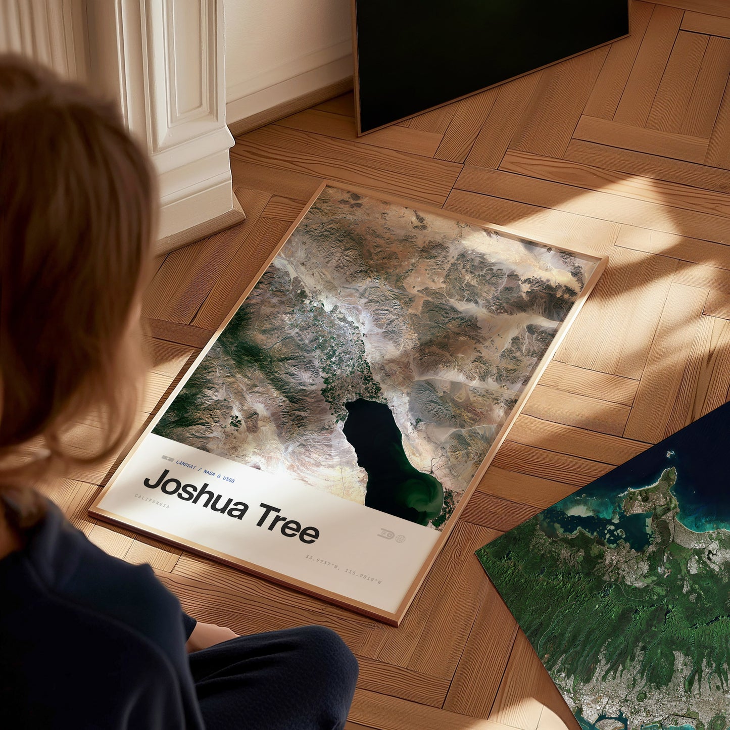 A person sits on a wooden floor, gazing at a framed poster of Joshua Tree, showcasing a detailed aerial view of the landscape, including mountains and a lake. Sunlight casts soft shadows, enhancing the textures of the art and the floor.