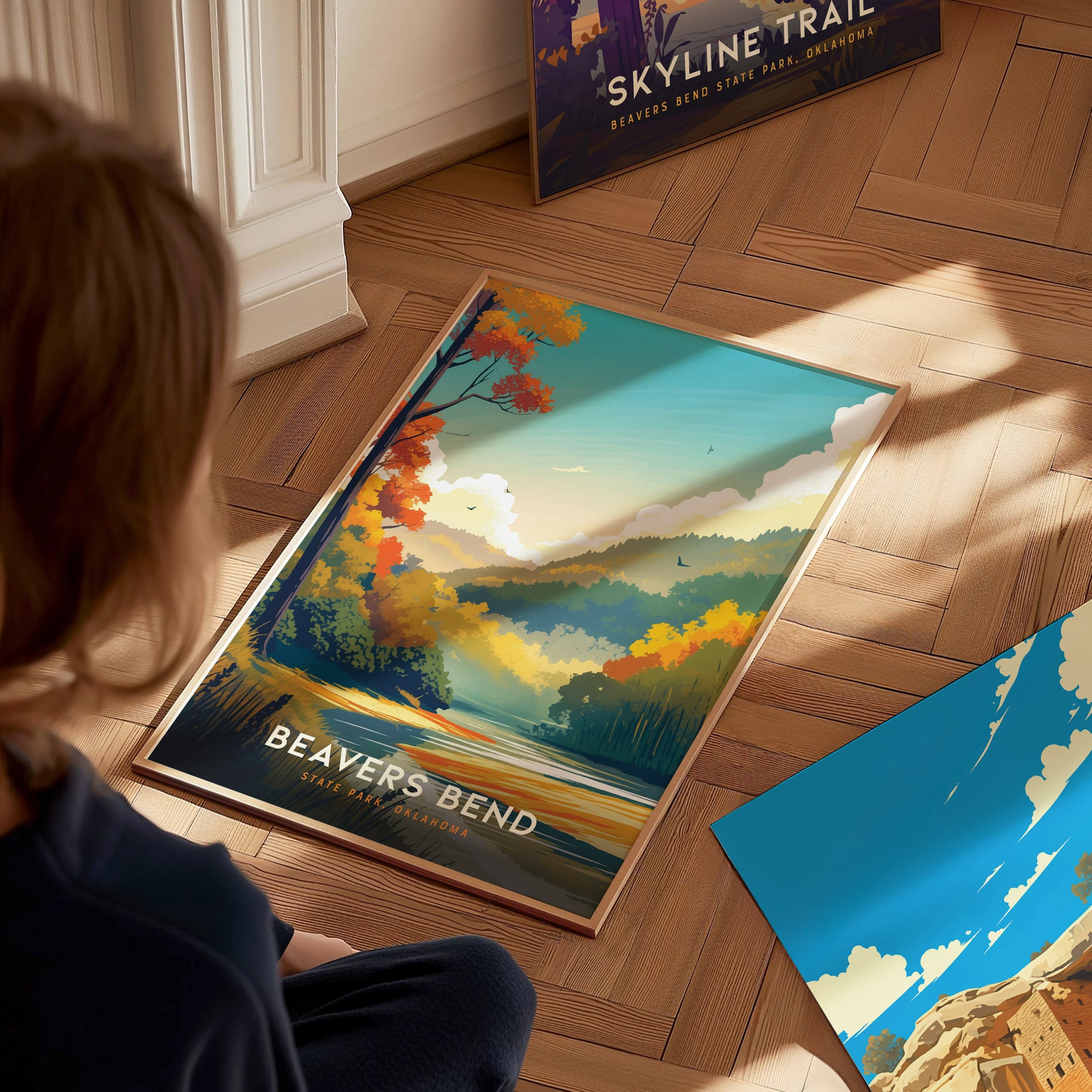 A person looks at a vibrant Beavers Bend State Park poster on a wooden floor, showcasing colorful trees against a scenic mountain backdrop. Other posters are partially visible nearby.