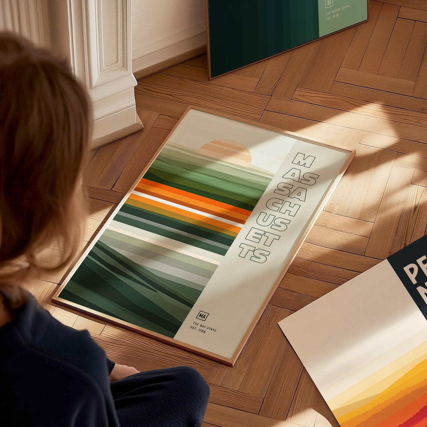 A person looks at a Massachusetts-themed poster featuring colorful horizontal stripes and bold text on a wooden floor. The poster is framed and partially illuminated by sunlight.