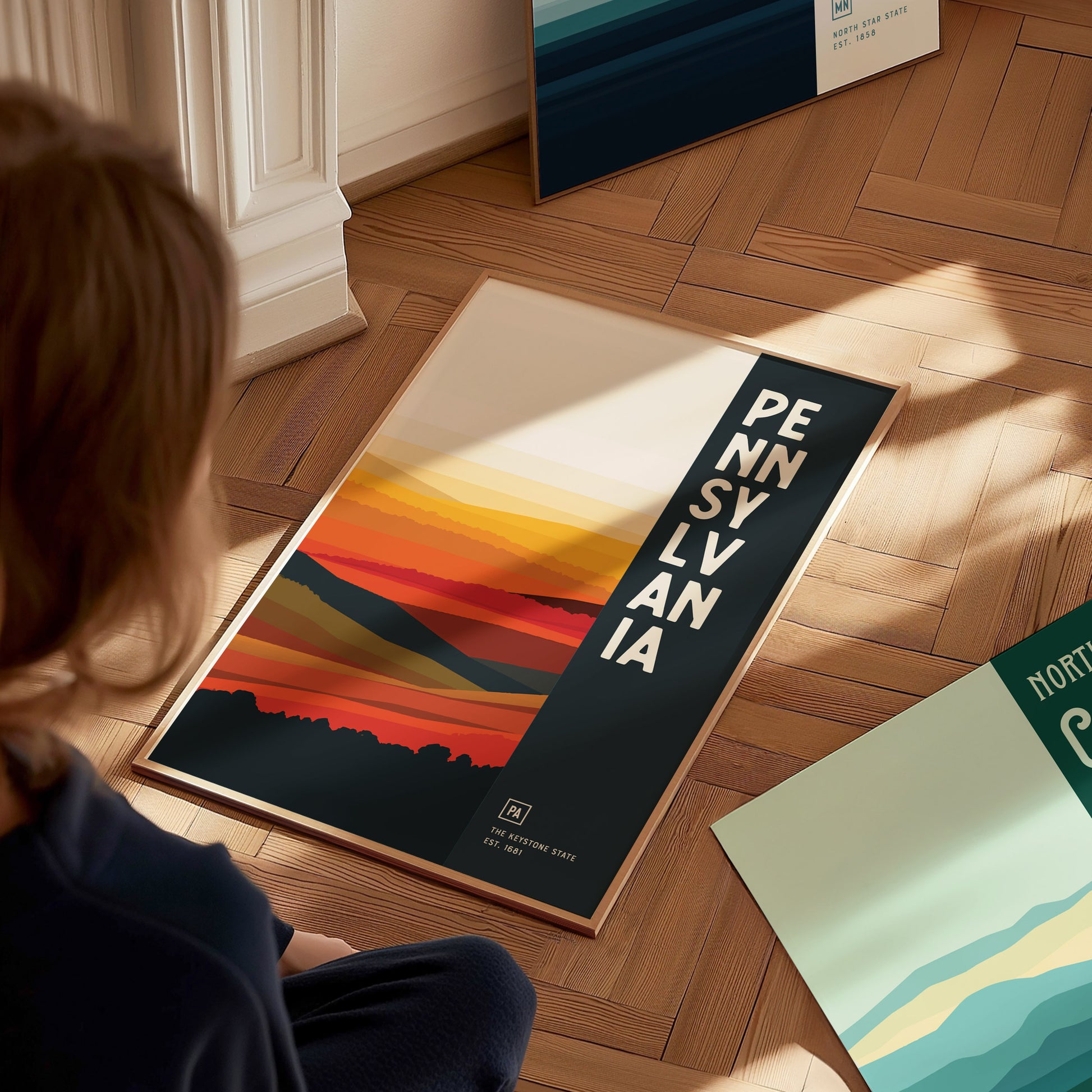 A person sits on a wooden floor, looking at a Pennsylvania poster with abstract, colorful landscape design and bold vertical text. Nearby is another partially visible poster.