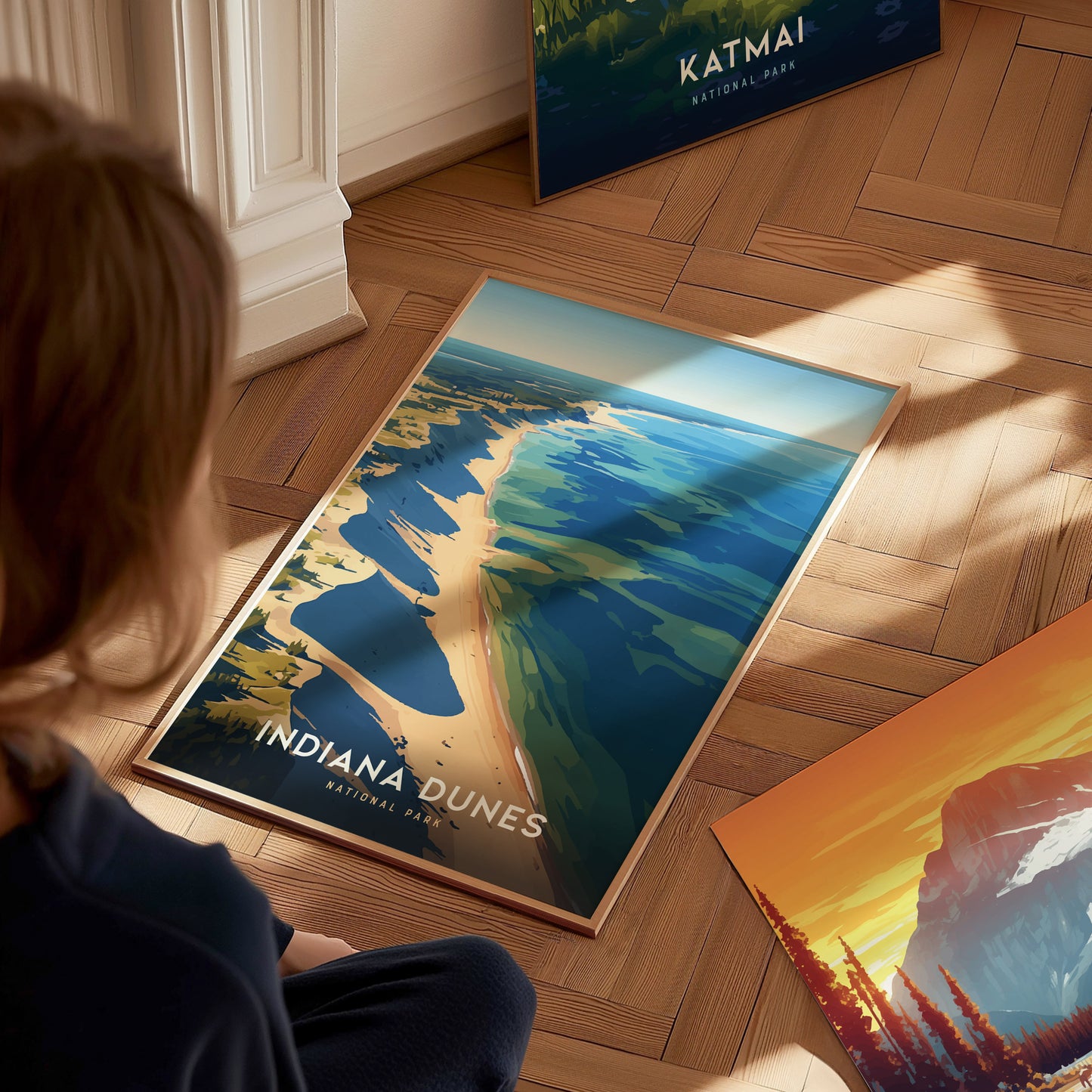 A person looks at a colorful Indiana Dunes National Park poster on a wooden floor, depicting a scenic shoreline and dune landscape. Other posters are partially visible nearby.