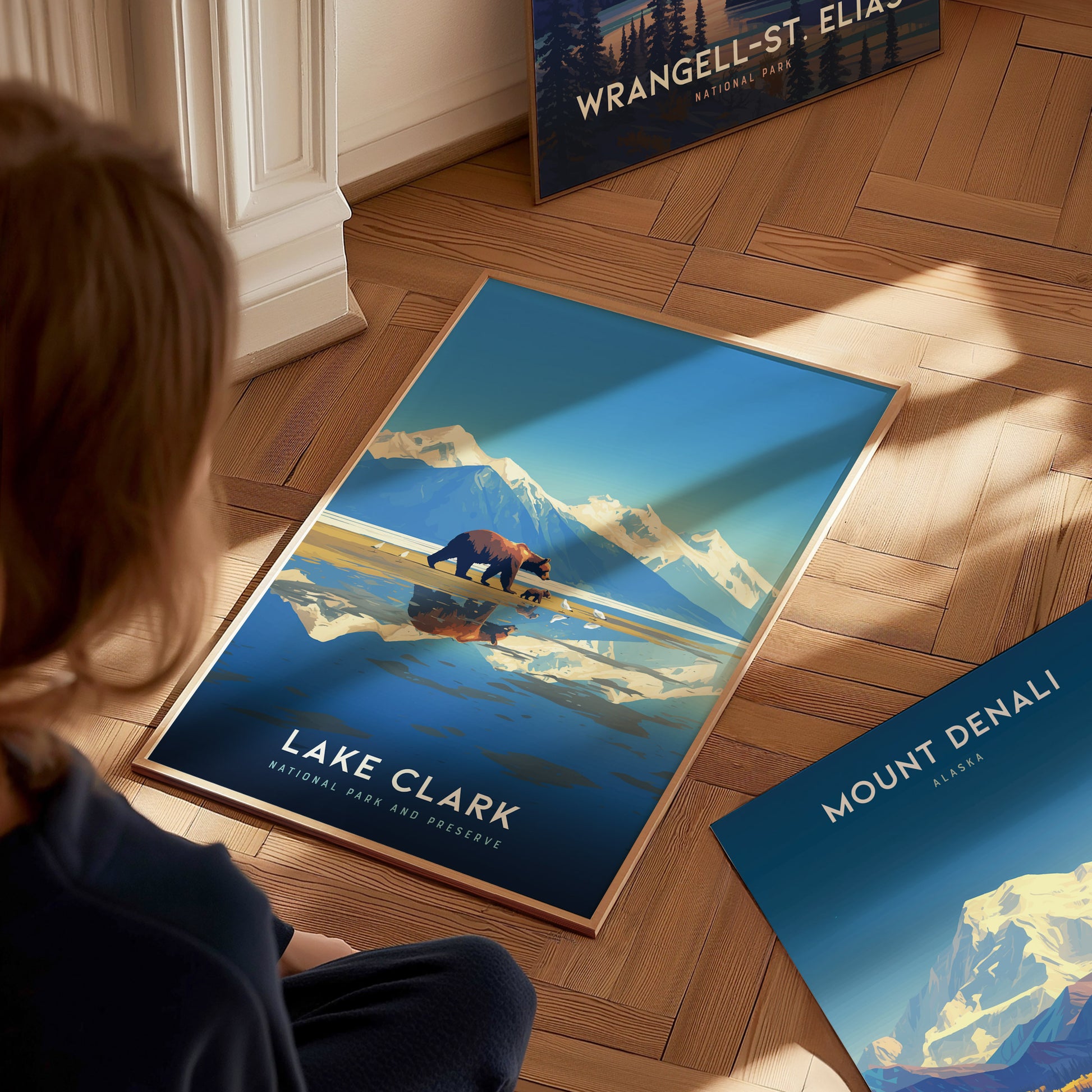 A person sits on a wooden floor admiring a Lake Clark National Park poster, featuring a bear against a backdrop of mountains and a reflective lake.