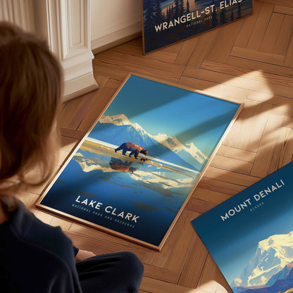 A person sits on a wooden floor admiring a Lake Clark National Park poster, featuring a bear against a backdrop of mountains and a reflective lake.