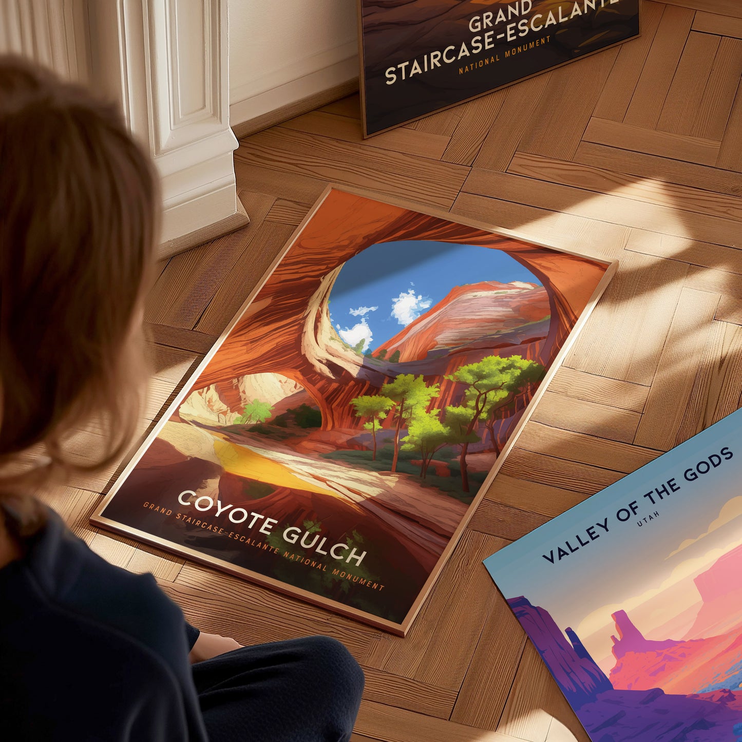 A person looks at a colorful poster of Coyote Gulch, depicting vibrant canyon walls and trees, placed on a wooden floor. Nearby, other posters are partially visible.