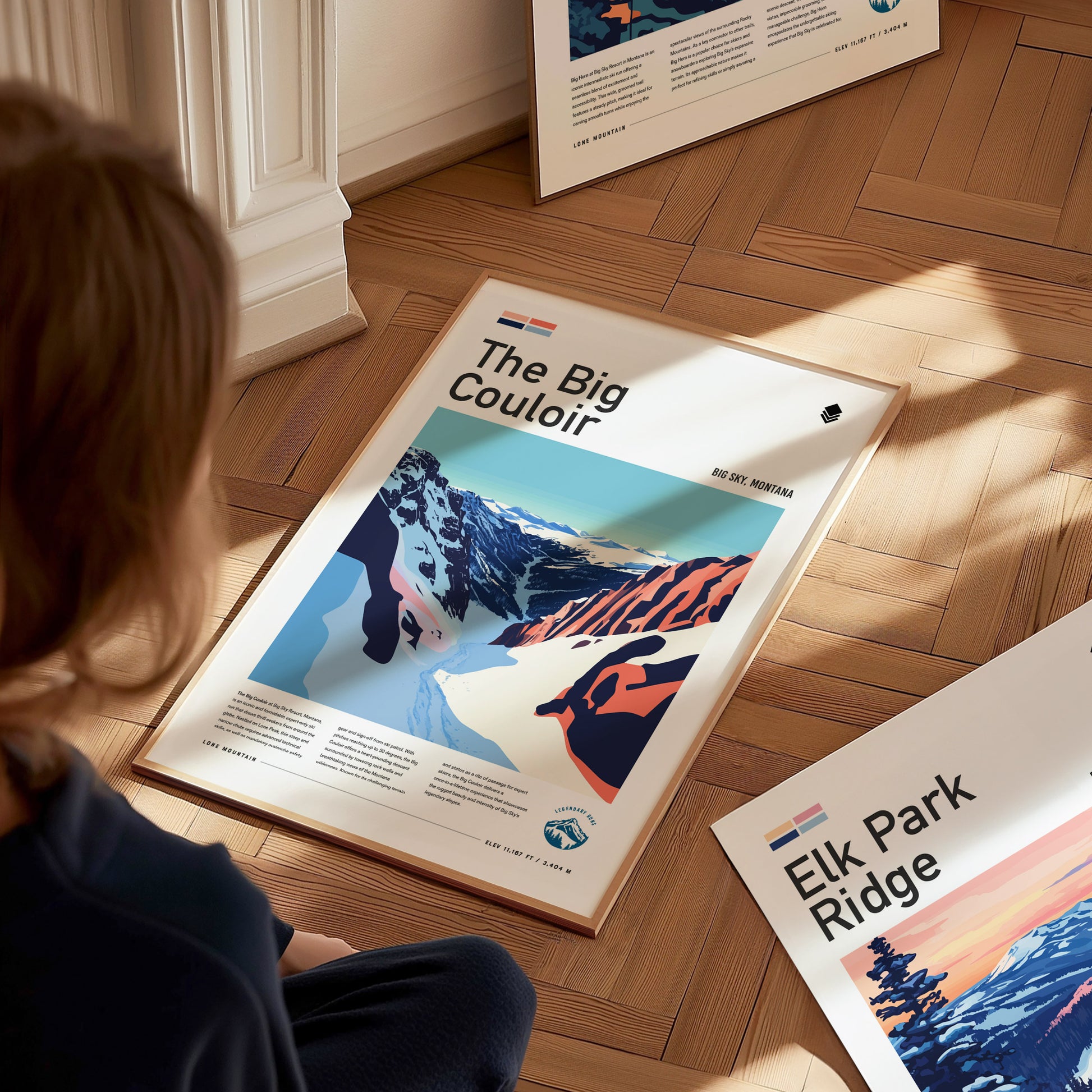 A person sits on a wooden floor looking at a framed poster titled The Big Couloir, featuring an abstract mountain landscape. Another poster titled Elk Park Ridge is nearby.