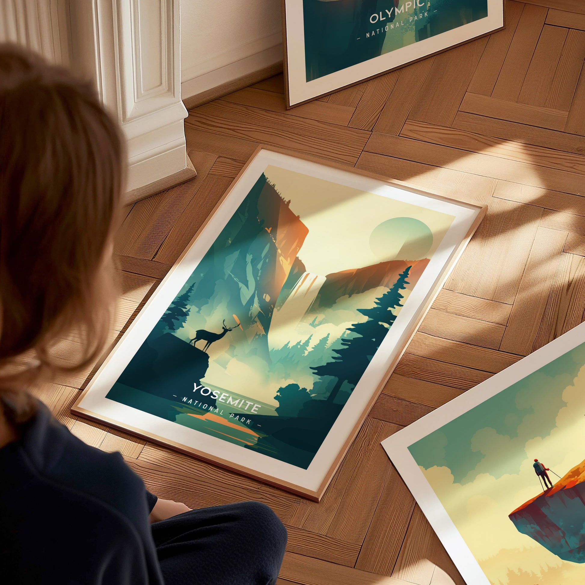 A person sits on a wooden floor observing a framed Yosemite National Park poster featuring a minimalist design of cliffs and pine trees. Another framed poster is partially visible nearby.