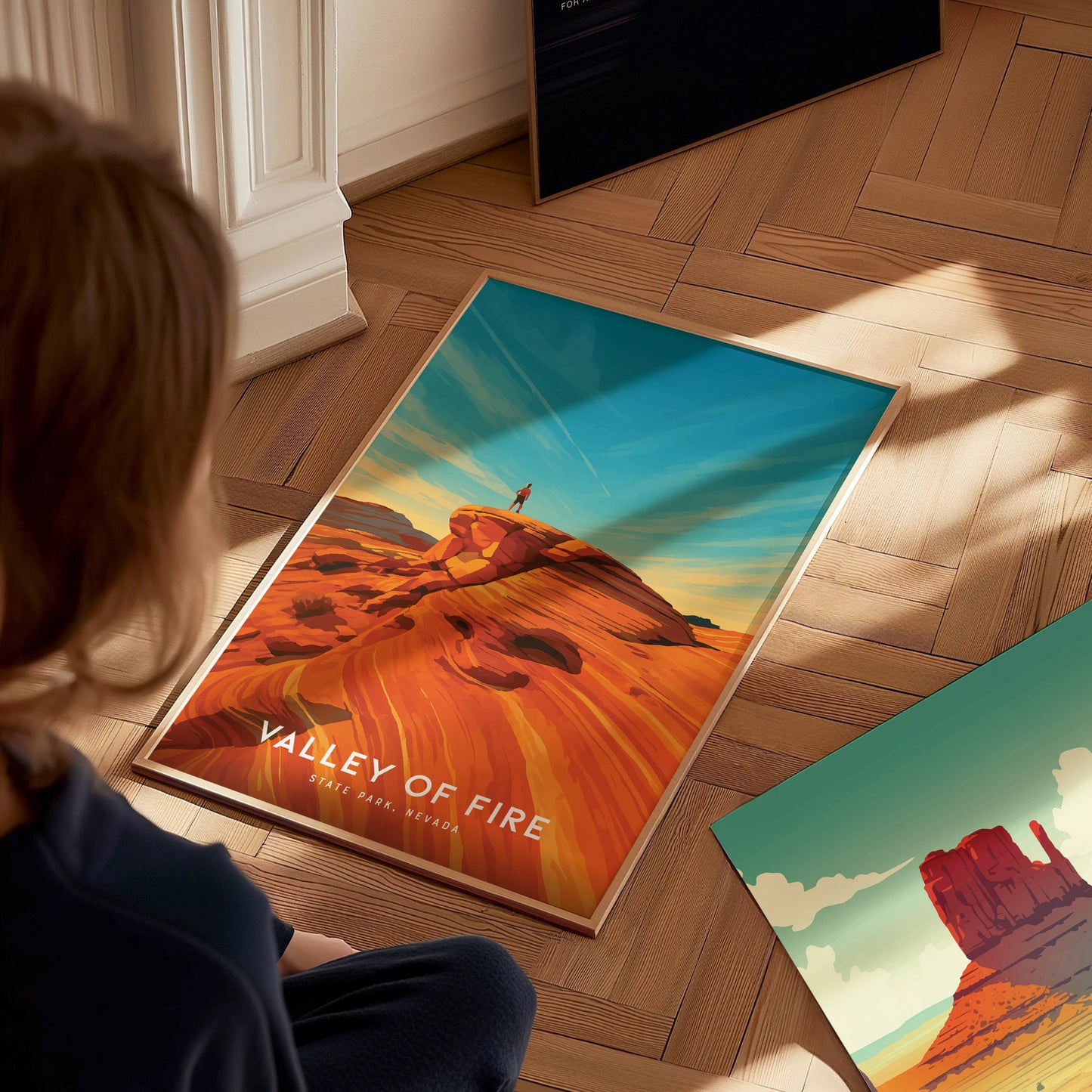 A person sits on a wooden floor, looking at two colorful travel prints. One print features a vibrant landscape titled Valley of Fire, showcasing striking red rock formations under a blue sky.