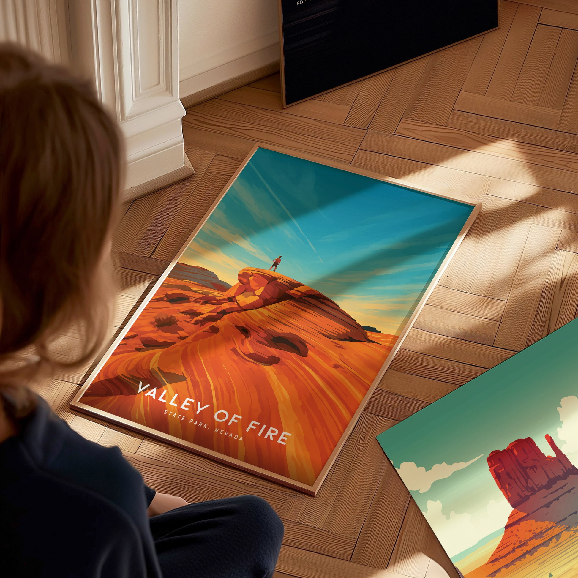 A person sits on a wooden floor, looking at two colorful travel prints. One print features a vibrant landscape titled Valley of Fire, showcasing striking red rock formations under a blue sky.