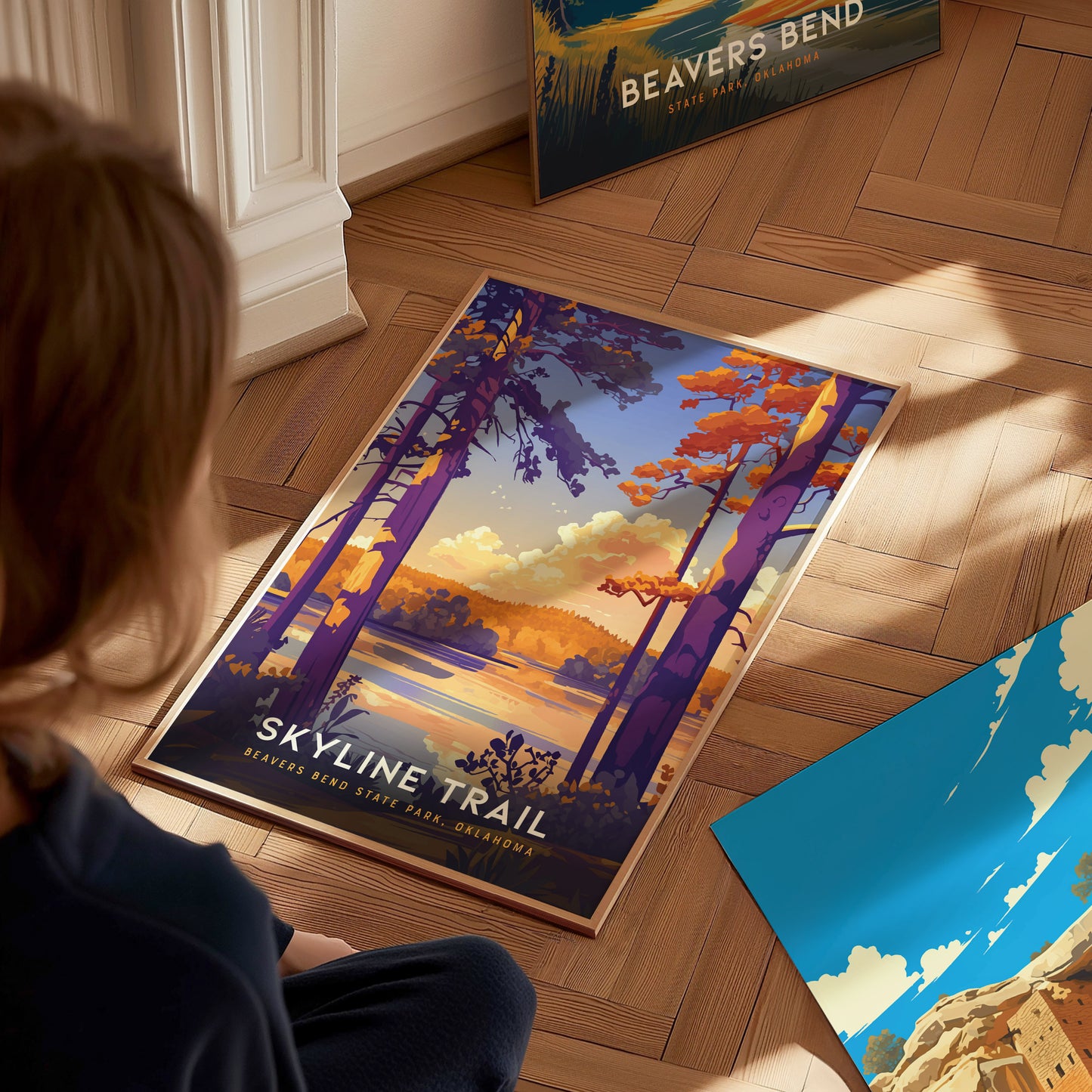 A person looks at a colorful Skyline Trail poster featuring vibrant trees and a scenic landscape. Another Beavers Bend poster leans against the wall on a wooden floor.