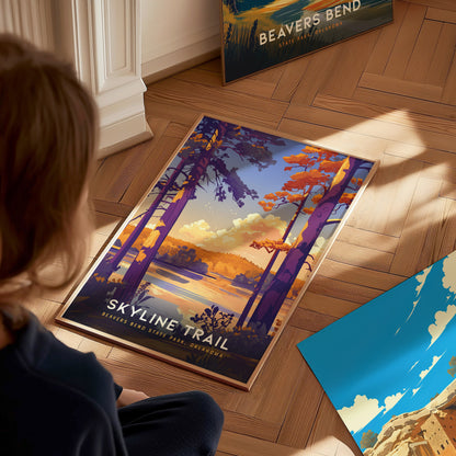 A person looks at a colorful Skyline Trail poster featuring vibrant trees and a scenic landscape. Another Beavers Bend poster leans against the wall on a wooden floor.