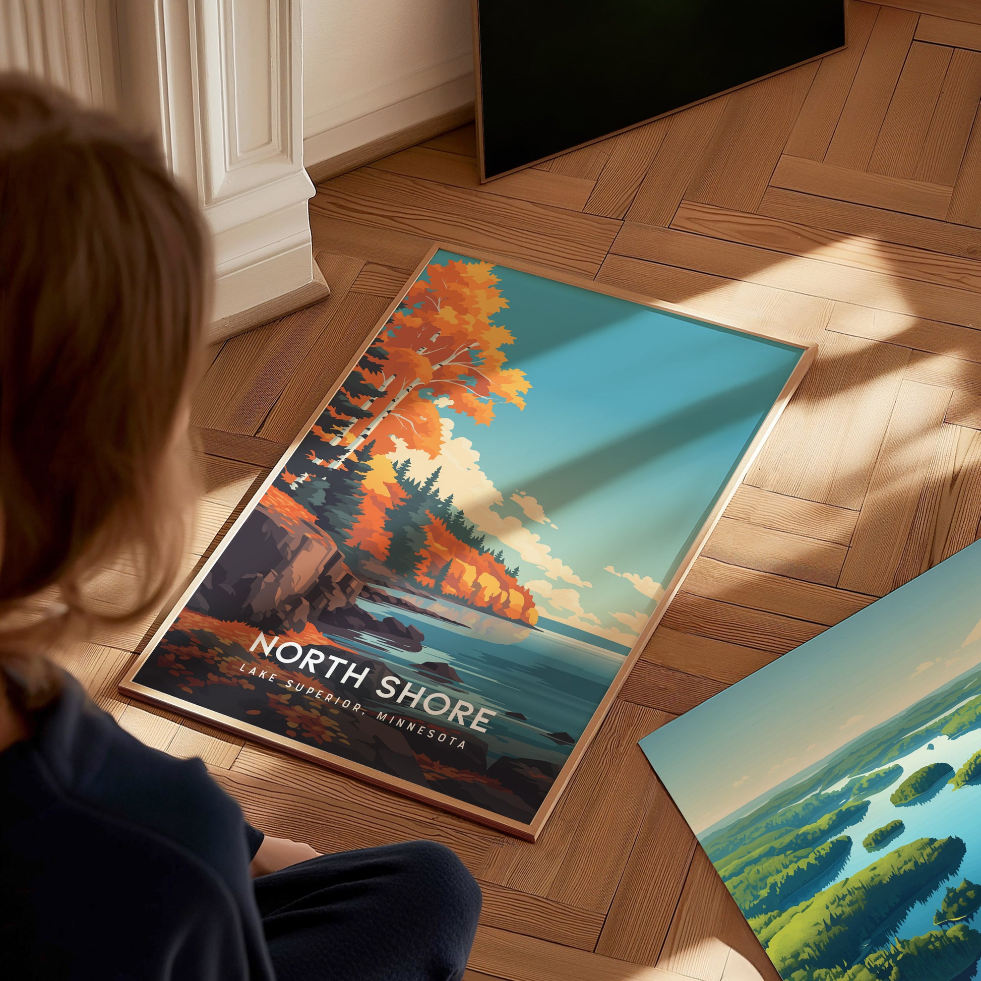 A child sits on a wooden floor, gazing at a colorful poster featuring the North Shore of Lake Superior.