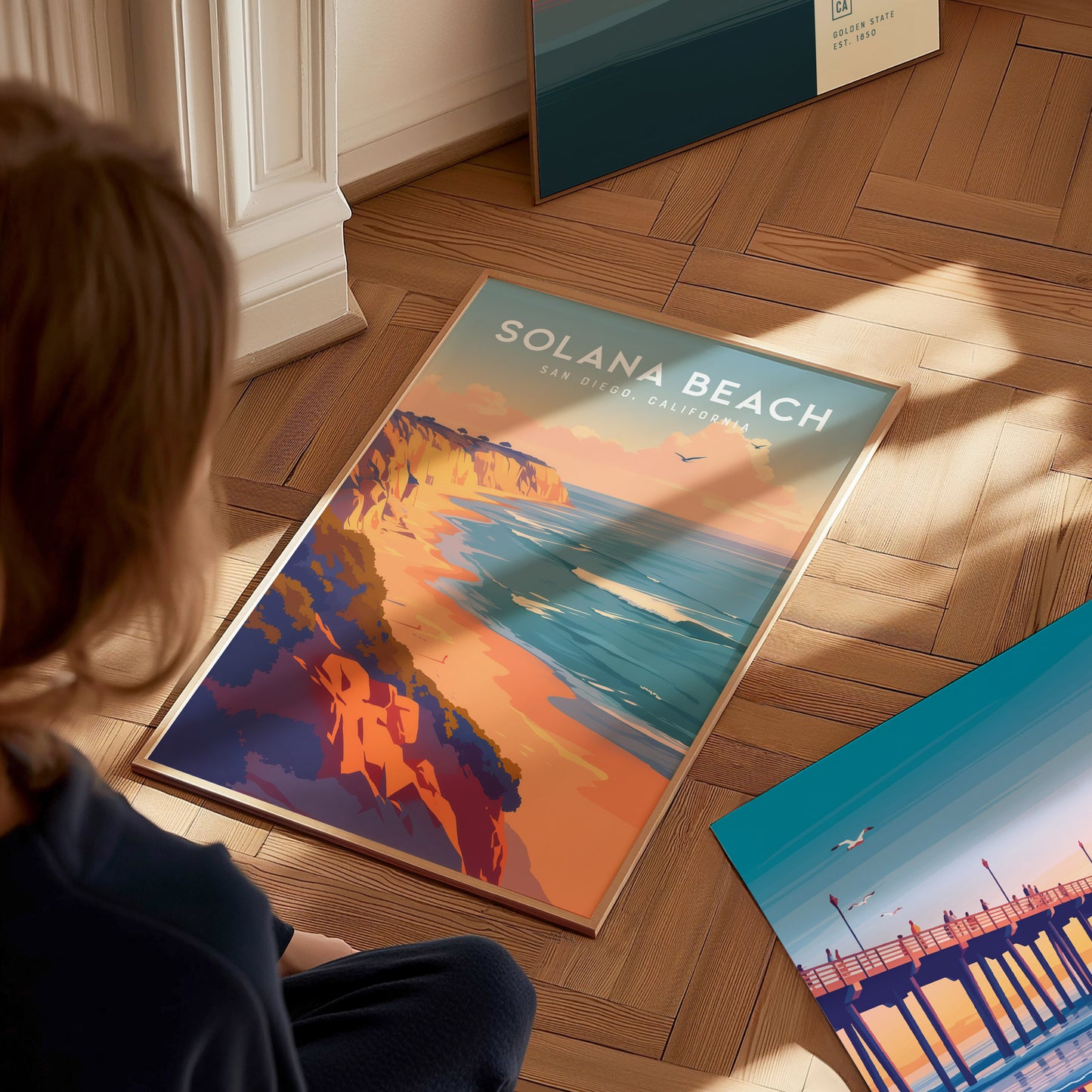 A person sits on a wooden floor, looking at a vibrant Solana Beach poster depicting coastal bluffs and ocean waves. The sunlight casts soft shadows across the scene.