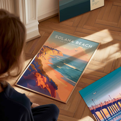 A person sits on a wooden floor, looking at a vibrant Solana Beach poster depicting coastal bluffs and ocean waves. The sunlight casts soft shadows across the scene.