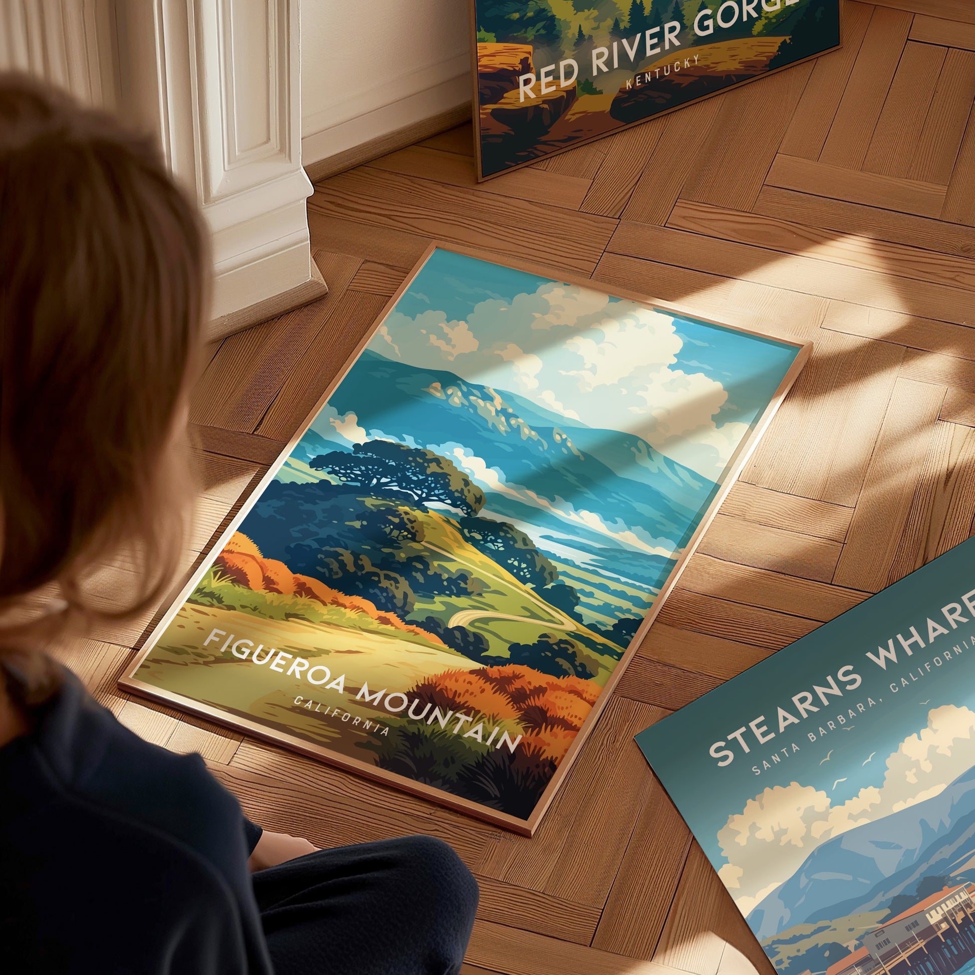 A person sits on a wooden floor, looking at a colorful poster featuring Figueroa Mountain in California. The illustration showcases vibrant rolling hills adorned with golden wildflowers, under a bright blue sky.