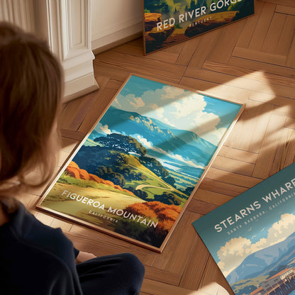 A person sits on a wooden floor, looking at a colorful poster featuring Figueroa Mountain in California. The illustration showcases vibrant rolling hills adorned with golden wildflowers, under a bright blue sky.