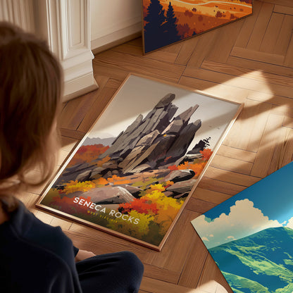 A person sits on a wooden floor, looking at a framed Seneca Rocks poster featuring a colorful autumn landscape with jagged rock formations. Other artworks are partially visible nearby.