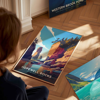 A person sits on a wooden floor, admiring a colorful Hopewell Rocks poster showcasing dramatic cliffs and vibrant skies. Other landscape posters are visible nearby.