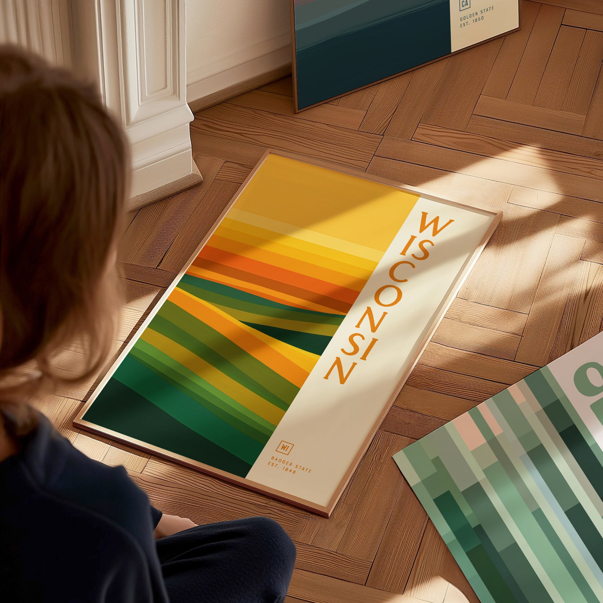 A person sits on the floor gazing at a Wisconsin poster featuring abstract green and gold waves. The poster rests on a wooden floor, capturing warm sunlight.