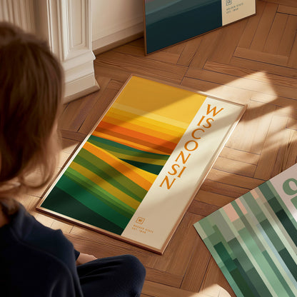 A person sits on the floor gazing at a Wisconsin poster featuring abstract green and gold waves. The poster rests on a wooden floor, capturing warm sunlight.