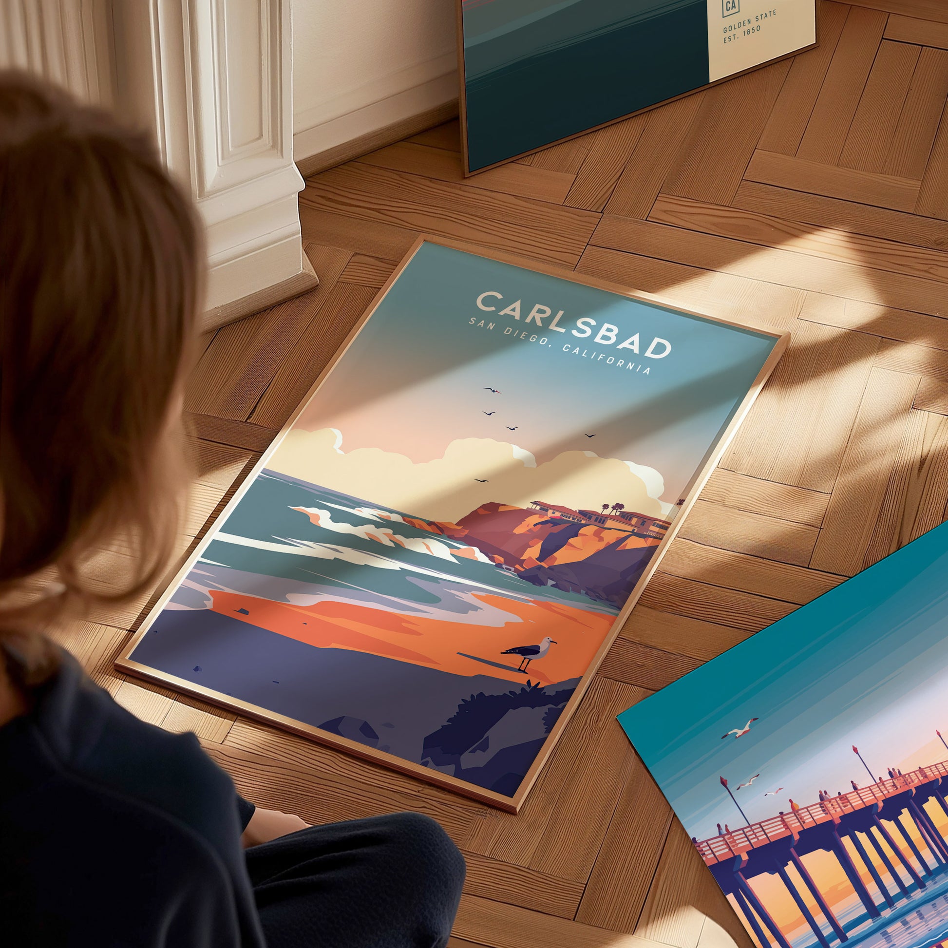 A person sits on a wooden floor, looking at a colorful poster of Carlsbad, San Diego, featuring cliffs and the ocean. Another ocean-themed print is nearby.