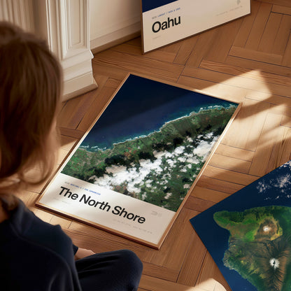 A child sits on a wooden floor, gazing at a framed travel poster featuring a satellite view of Oahus North Shore.