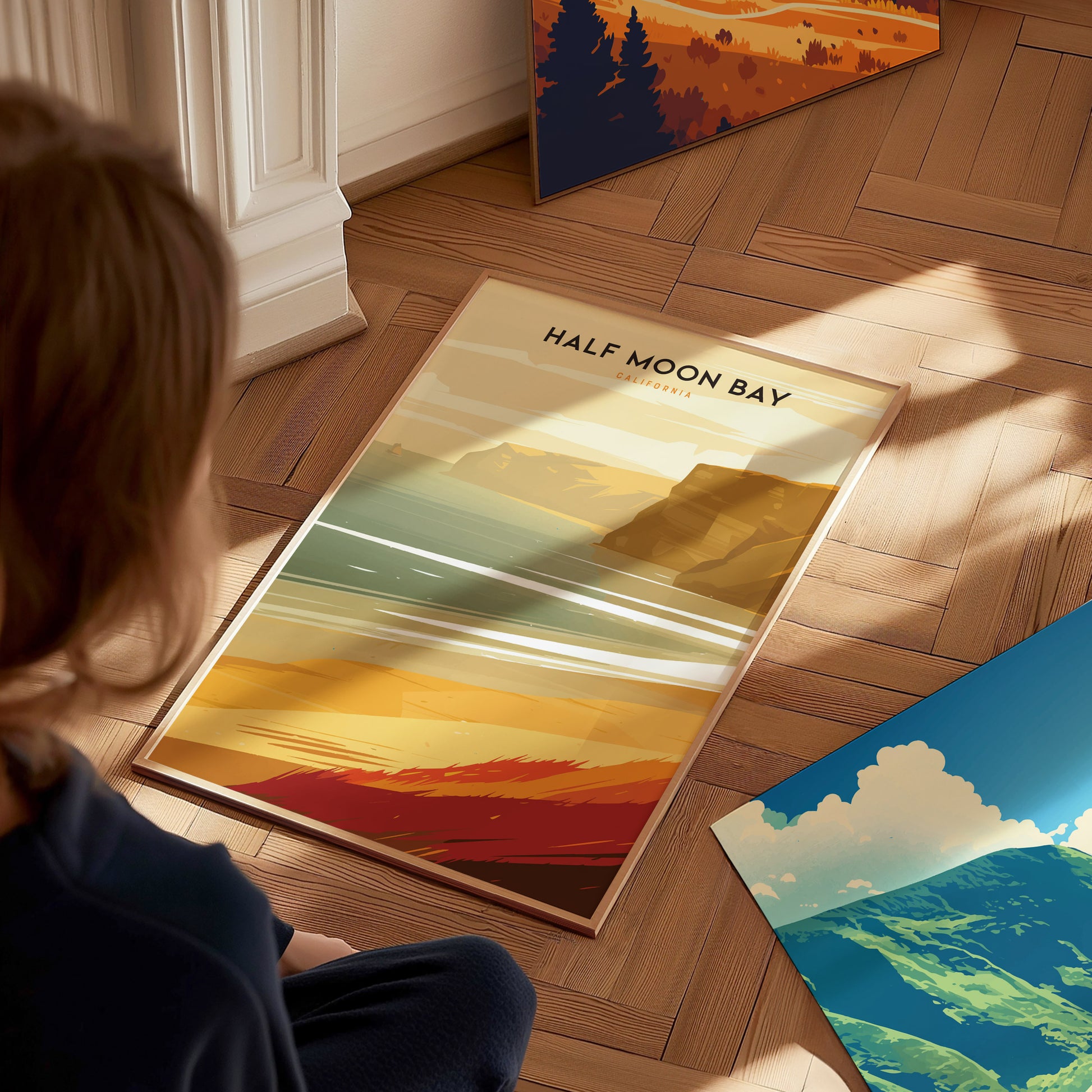 A person is sitting on a wooden floor, looking at a Half Moon Bay poster depicting a coastal scene with golden cliffs and ocean waves.