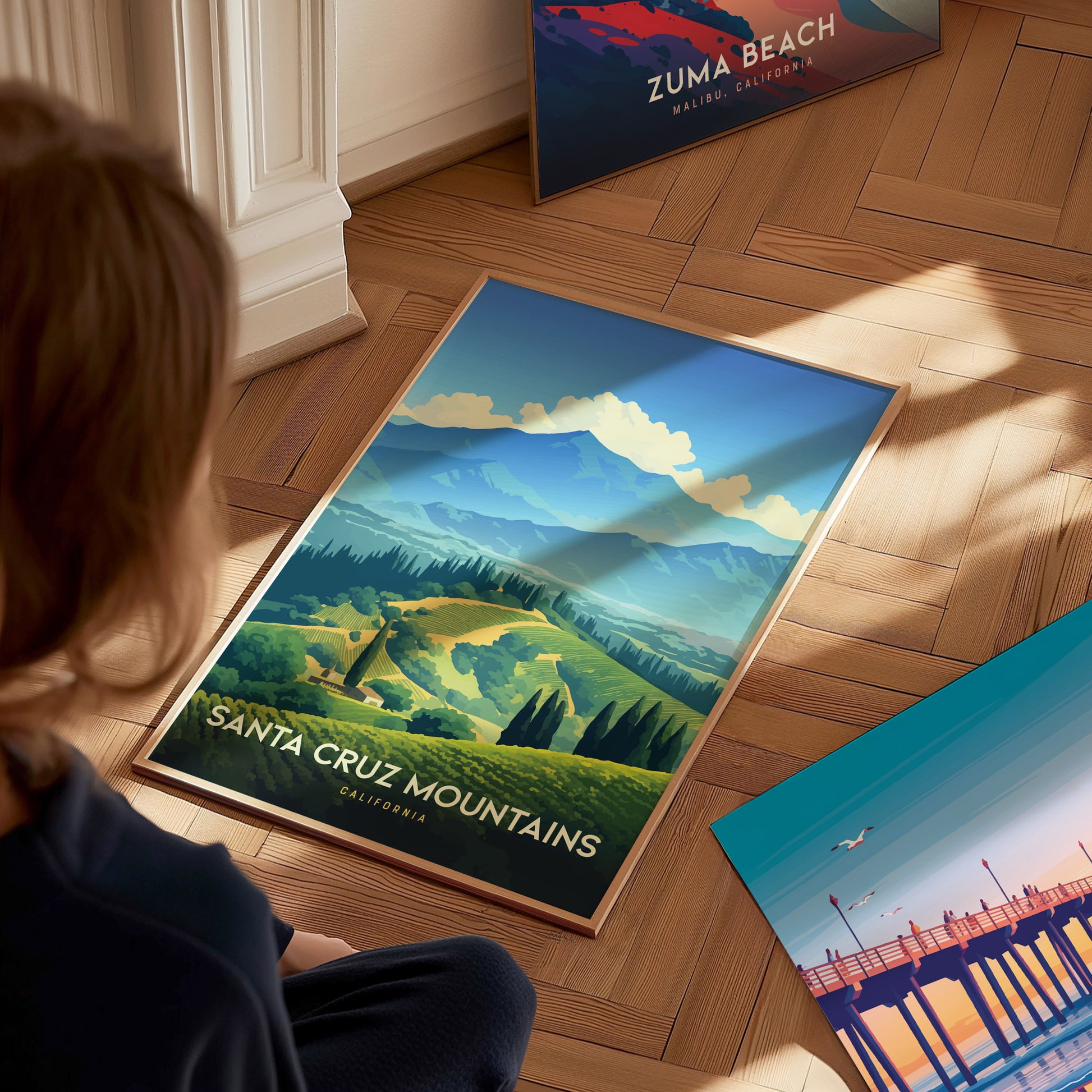 A person sits on a wooden floor, looking at a framed Santa Cruz Mountains poster featuring vibrant vineyard hills and mountains. Other posters are partially visible nearby.