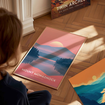 A person observes a framed Mount Monadnock poster depicting a colorful sunset over a mountain and lake, placed on a wooden floor with another landscape poster nearby.