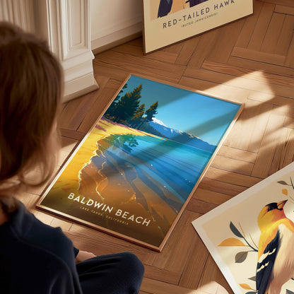 A person sits on a wooden floor, looking at a vibrant Baldwin Beach, Lake Tahoe poster depicting clear waters and a forested shoreline.