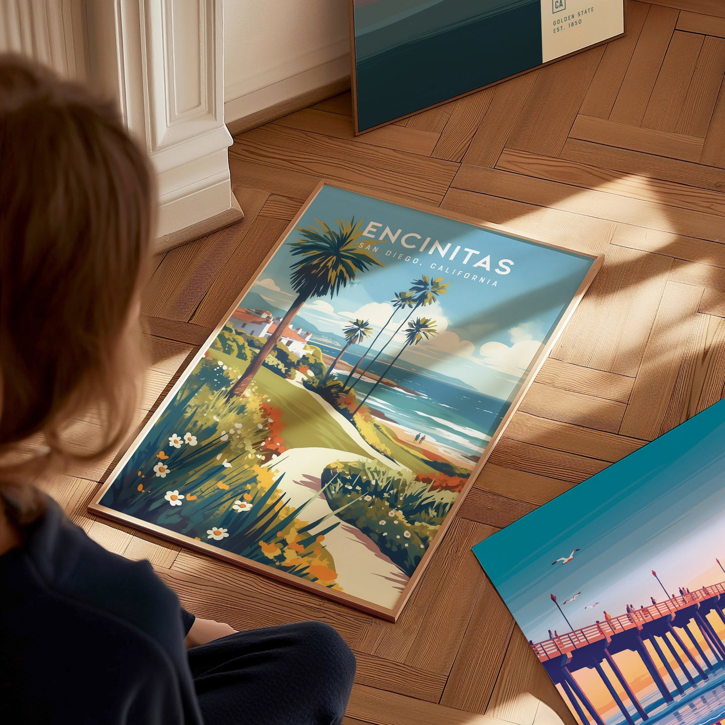 A person views a colorful poster of Encinitas, California, featuring palm trees, a coastal path, and ocean waves, placed on a wooden floor beside another vibrant artwork.