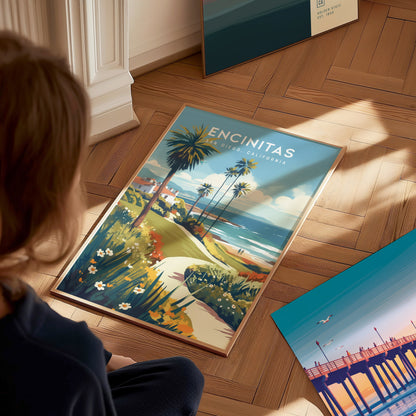 A person views a colorful poster of Encinitas, California, featuring palm trees, a coastal path, and ocean waves, placed on a wooden floor beside another vibrant artwork.