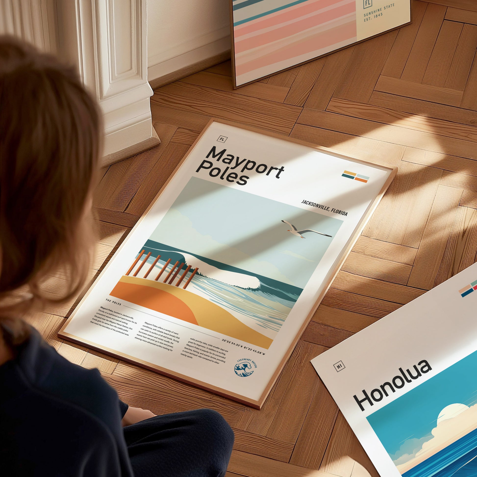 A person sits on a wooden floor, looking at a framed Mayport Poles poster featuring a beach scene with waves and a seagull. Other colorful posters are visible nearby.