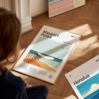 A person sits on a wooden floor, looking at a framed Mayport Poles poster featuring a beach scene with waves and a seagull. Other colorful posters are visible nearby.