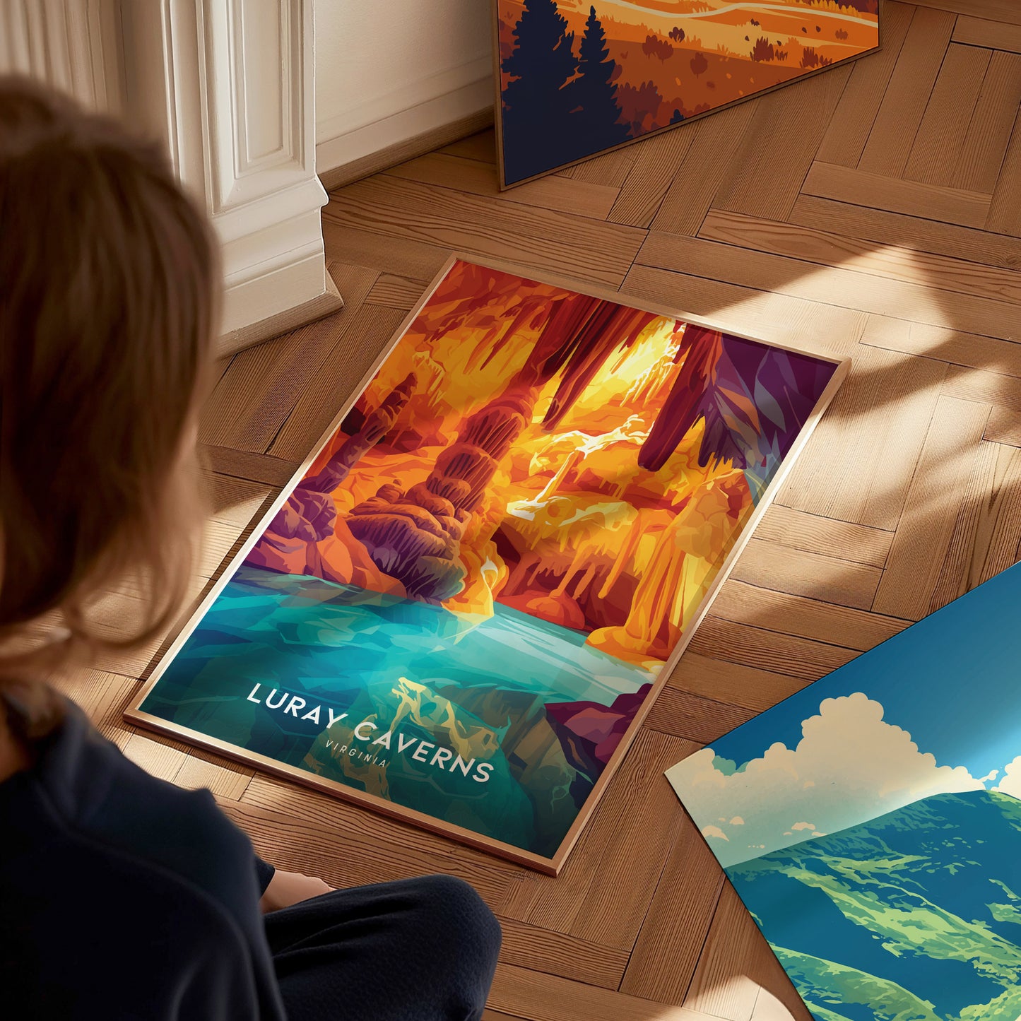 A person sits on a wooden floor, looking at a colorful Luray Caverns poster featuring vibrant cave formations and a turquoise pool, with additional artwork nearby.