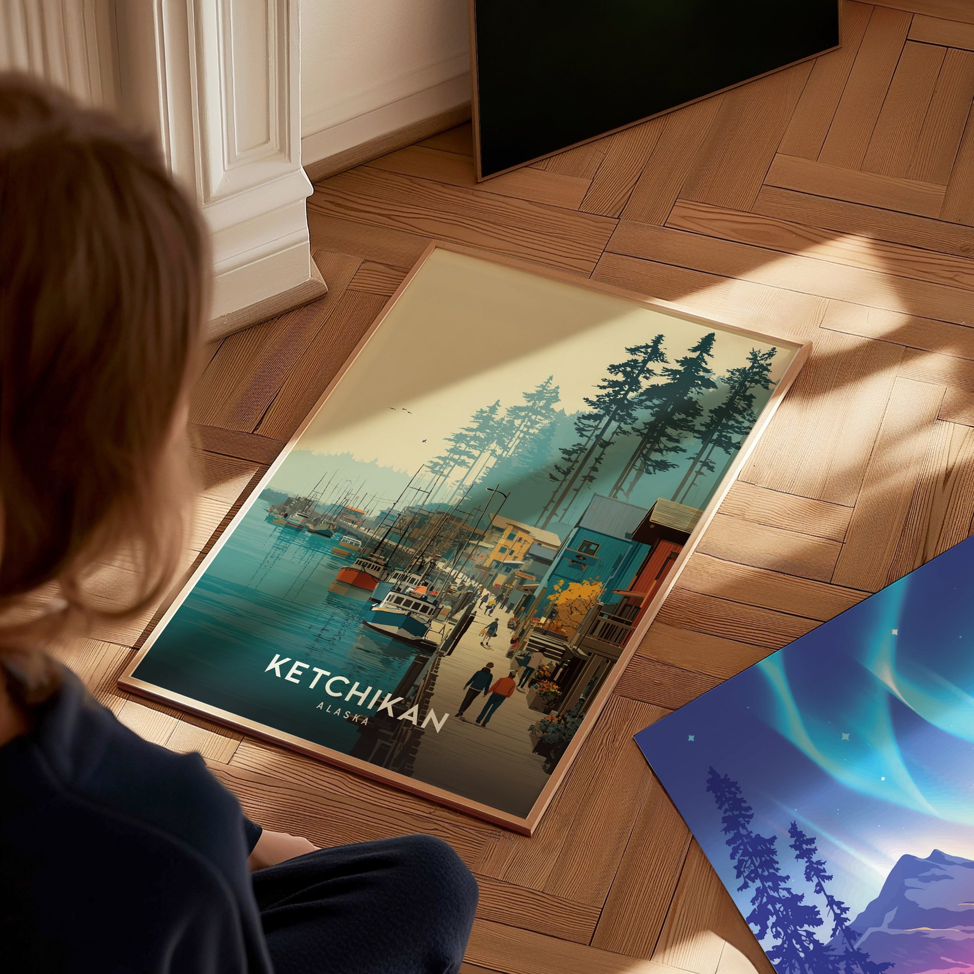 A person is sitting on a hardwood floor, gazing at a colorful art print featuring a scenic view of Ketchikan, Alaska, with boats in a harbor and tall trees in the background. The warm tones and detailed artwork create a vibrant atmosphere.