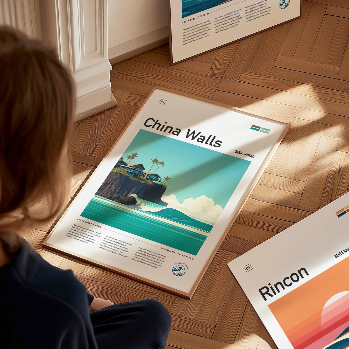 A person sits on a wooden floor, looking at a framed China Walls poster featuring a coastal scene with turquoise waves, cliffs, and palm trees. Additional posters are partially visible.