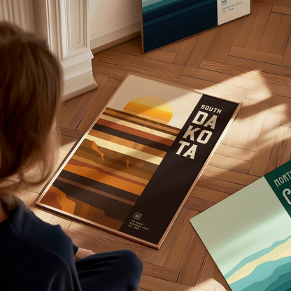 A person looks at a South Dakota poster featuring a minimalist geometric design with earthy tones and abstract landscape shapes on a wooden floor. Additional posters are partially visible.