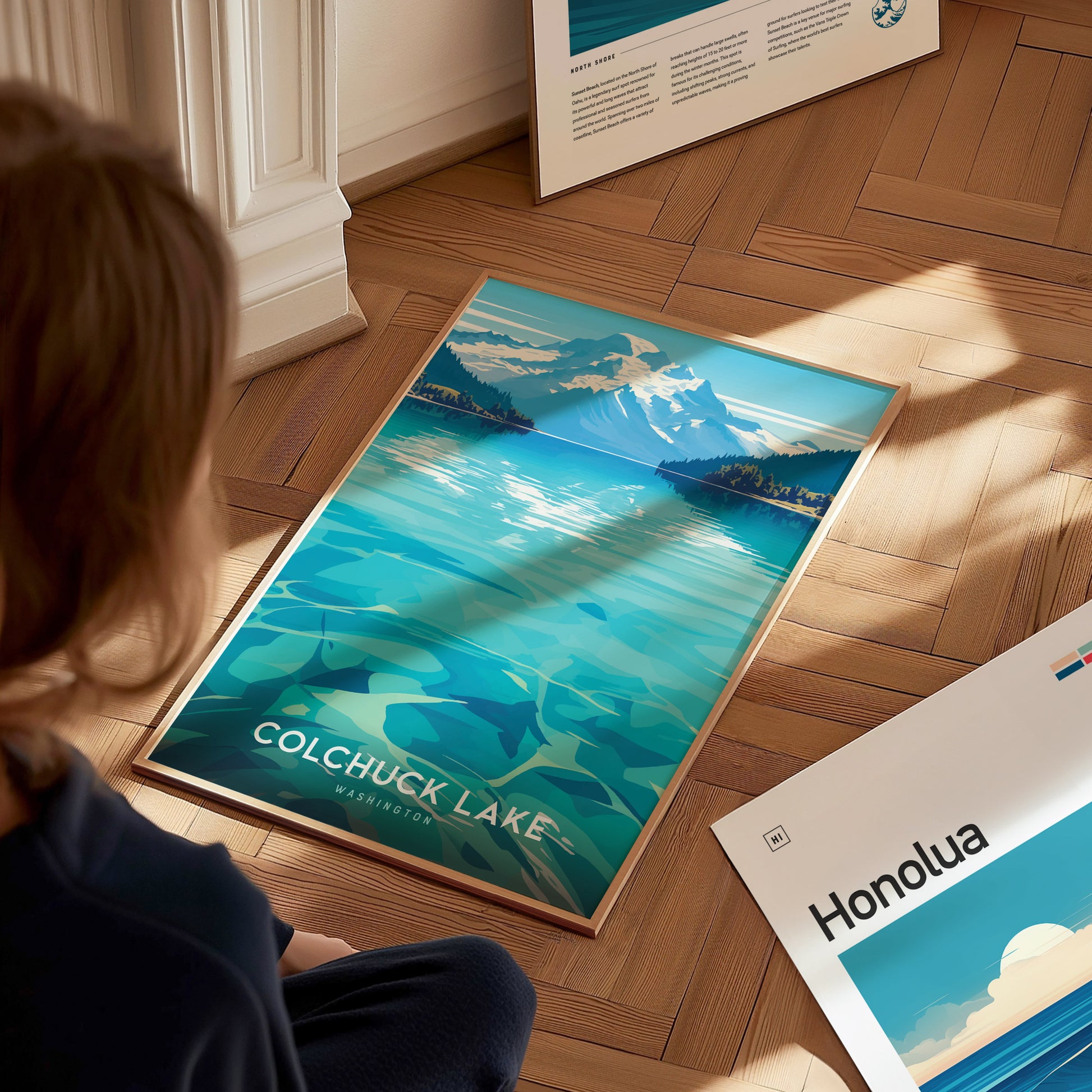 A person is looking at a framed Colchuck Lake poster on a wooden floor, depicting turquoise waters and mountains. Another poster titled Honolua is partially visible nearby.