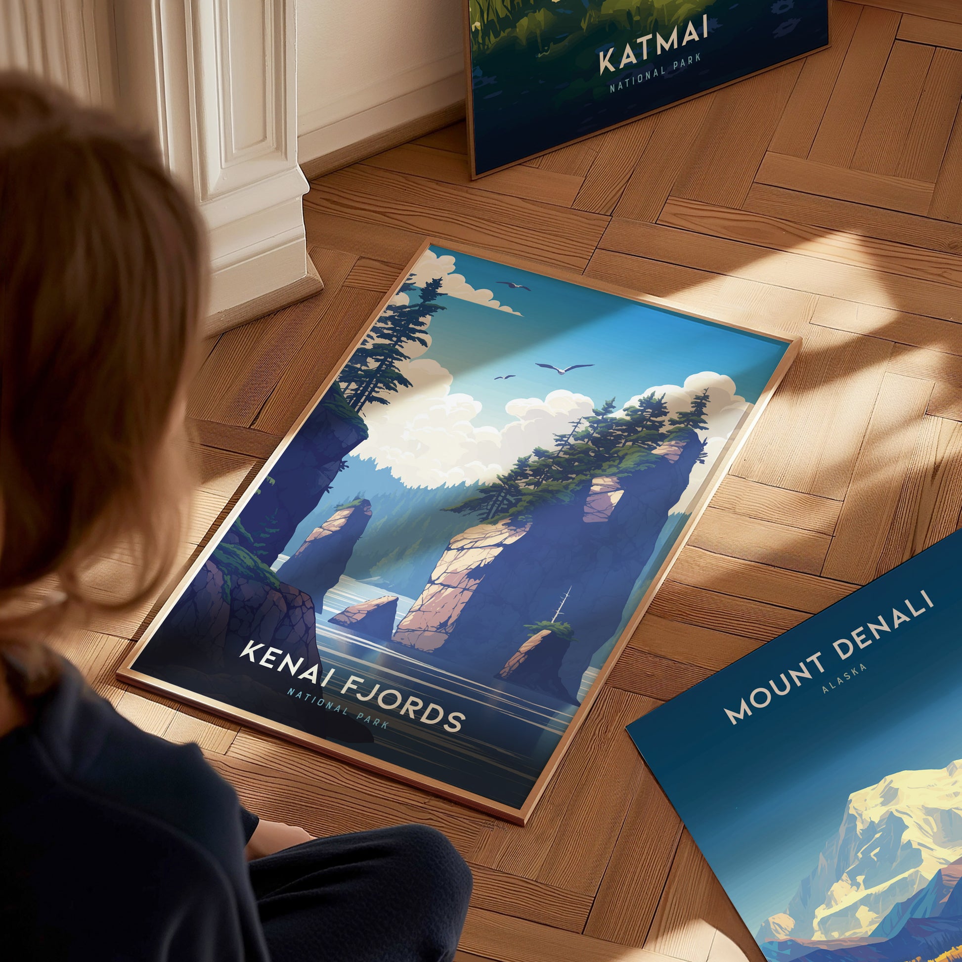A person views a Kenai Fjords National Park poster depicting rocky cliffs, trees, and a vibrant sky. Nearby, Katmai and Mount Denali posters rest on a wooden floor.