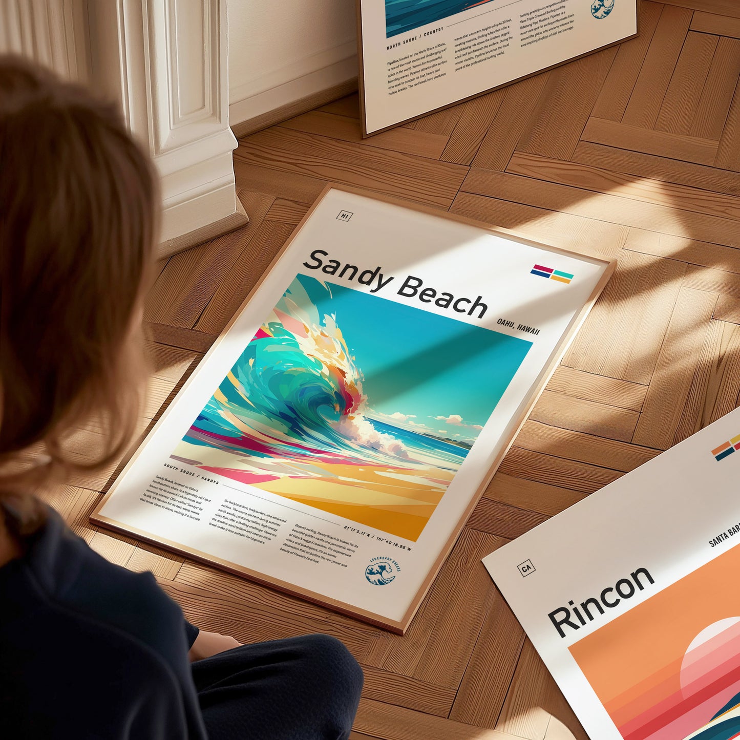A person is looking at a framed Sandy Beach poster on a wooden floor, featuring vibrant ocean waves and bright colors. Other framed posters are partially visible nearby.