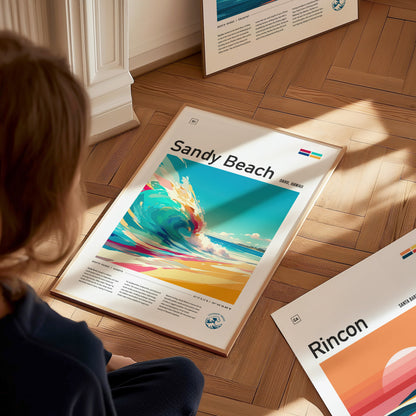 A person is looking at a framed Sandy Beach poster on a wooden floor, featuring vibrant ocean waves and bright colors. Other framed posters are partially visible nearby.
