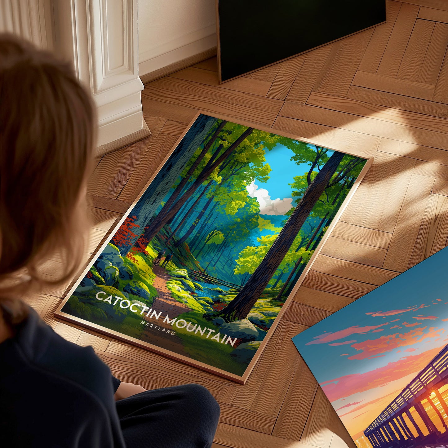 A person sits on a wooden floor, gazing at a colorful poster of Catoctin Mountain, Maryland, featuring a vibrant forest path surrounded by lush green trees and a bright blue sky.