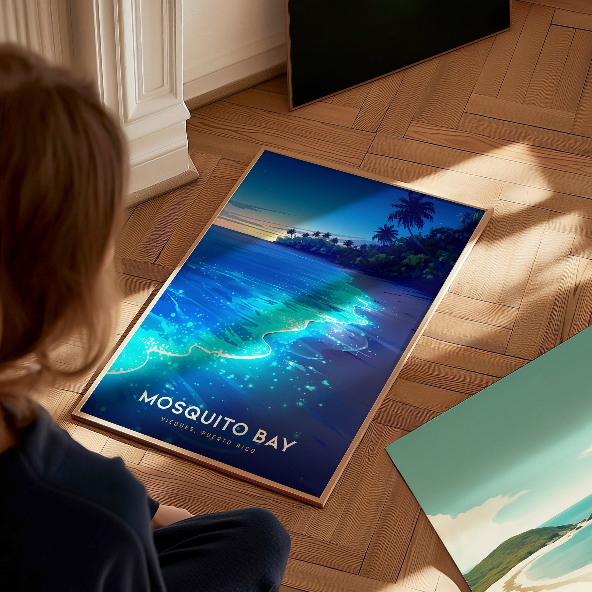 A person sits on a wooden floor, gazing at a vibrant poster featuring Mosquito Bay in Vieques, Puerto Rico. The artwork depicts glowing blue waters and palm trees under a serene sky, capturing the enchanting beauty of the bay.