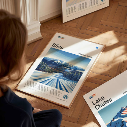 A person sits on a wooden floor viewing a framed Bliss ski run poster featuring Breckenridge, Colorado, depicting a snowy landscape with mountains and trees under a clear blue sky.