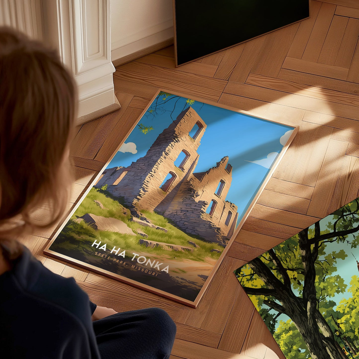 A person looks at a framed poster of Ha Ha Tonka State Park, featuring stone ruins under a blue sky, placed on a wooden floor. Sunlight casts soft shadows.