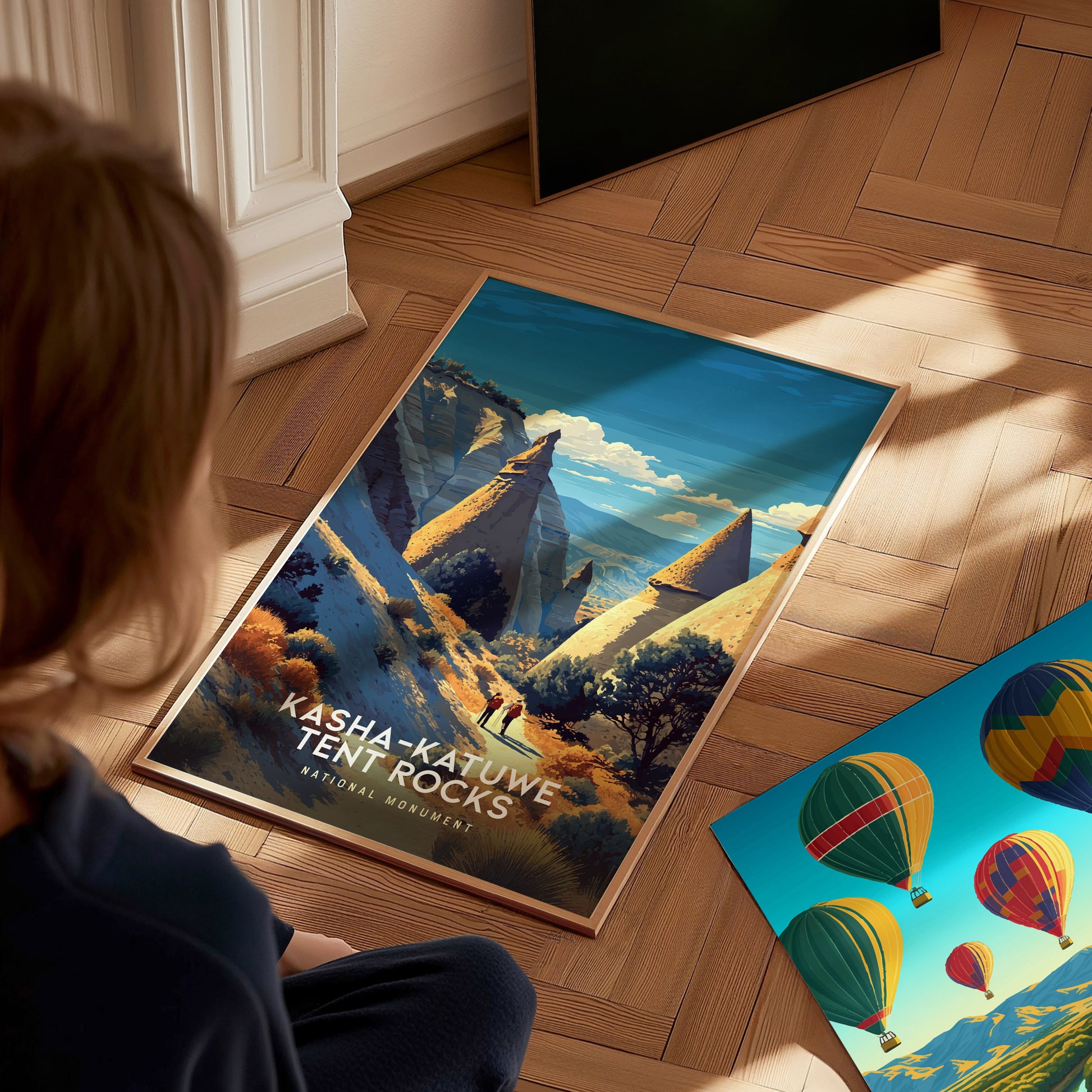 A person sits on a wooden floor, gazing at a vibrant vintage travel print depicting the Kasha-Katuwe Tent Rocks National Monument.