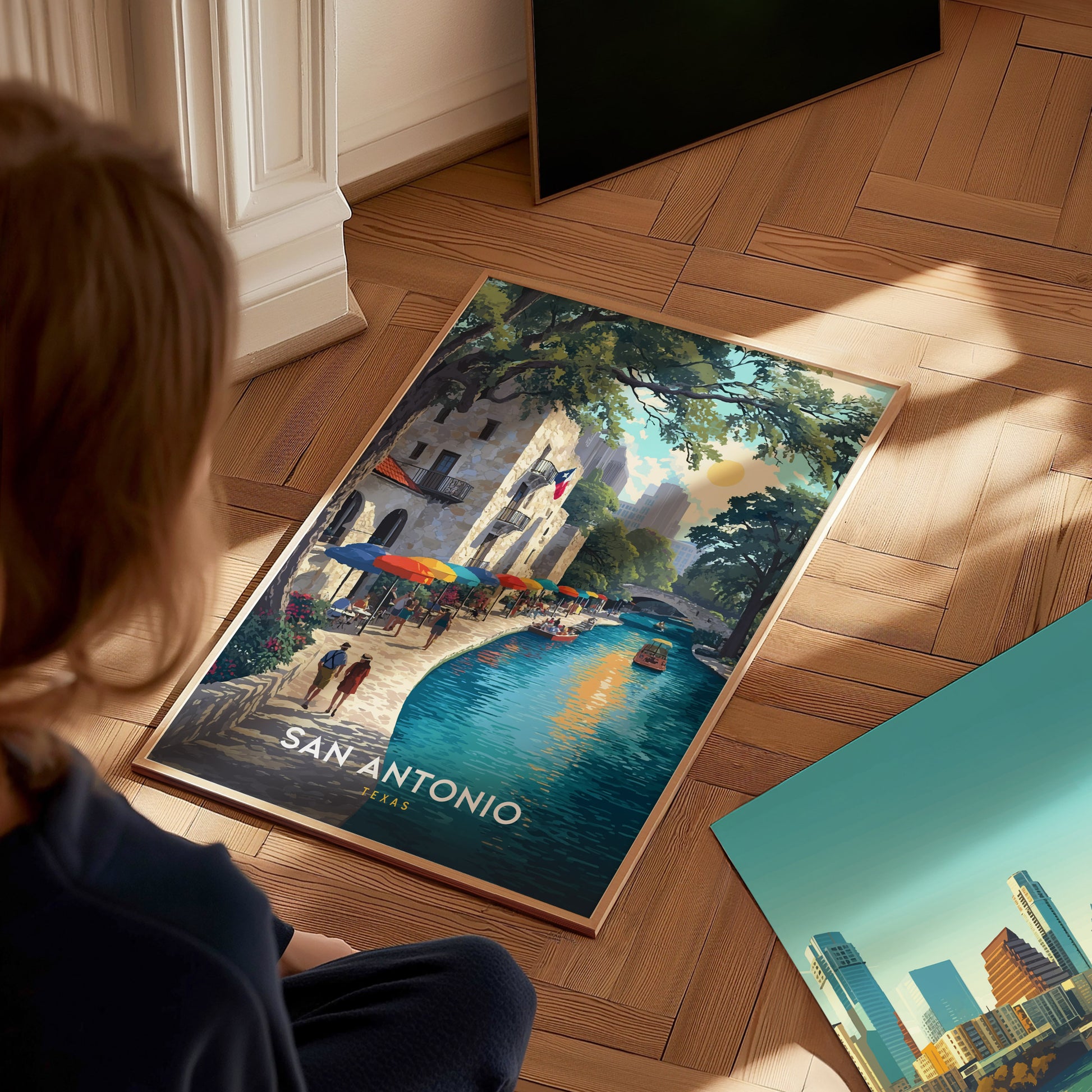A person looks at a vibrant poster on a wooden floor, depicting San Antonios River Walk with colorful umbrellas and a scenic river. Sunlight streams through a nearby window.